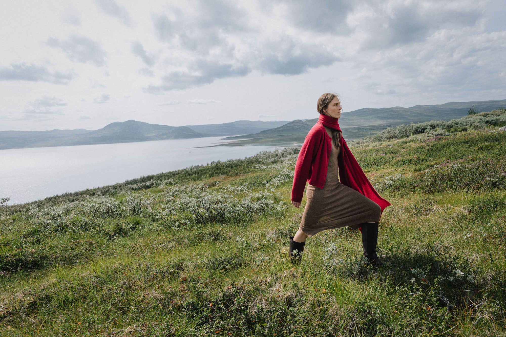 Person in a red cloak standing on a grassy hill with a lake and mountains in the background
