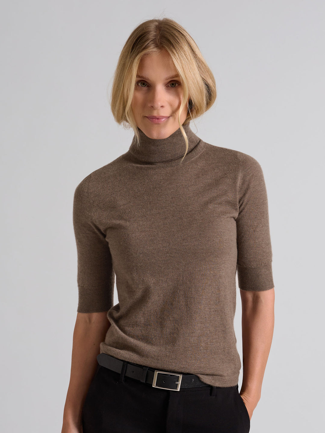 Woman wearing a brown turtleneck sweater against a gray background. Turtleneck cashmere t-shirt "Agnes" in 100% pure cashmere. Scandinavian design by Kashina. Color: Dark Toast.