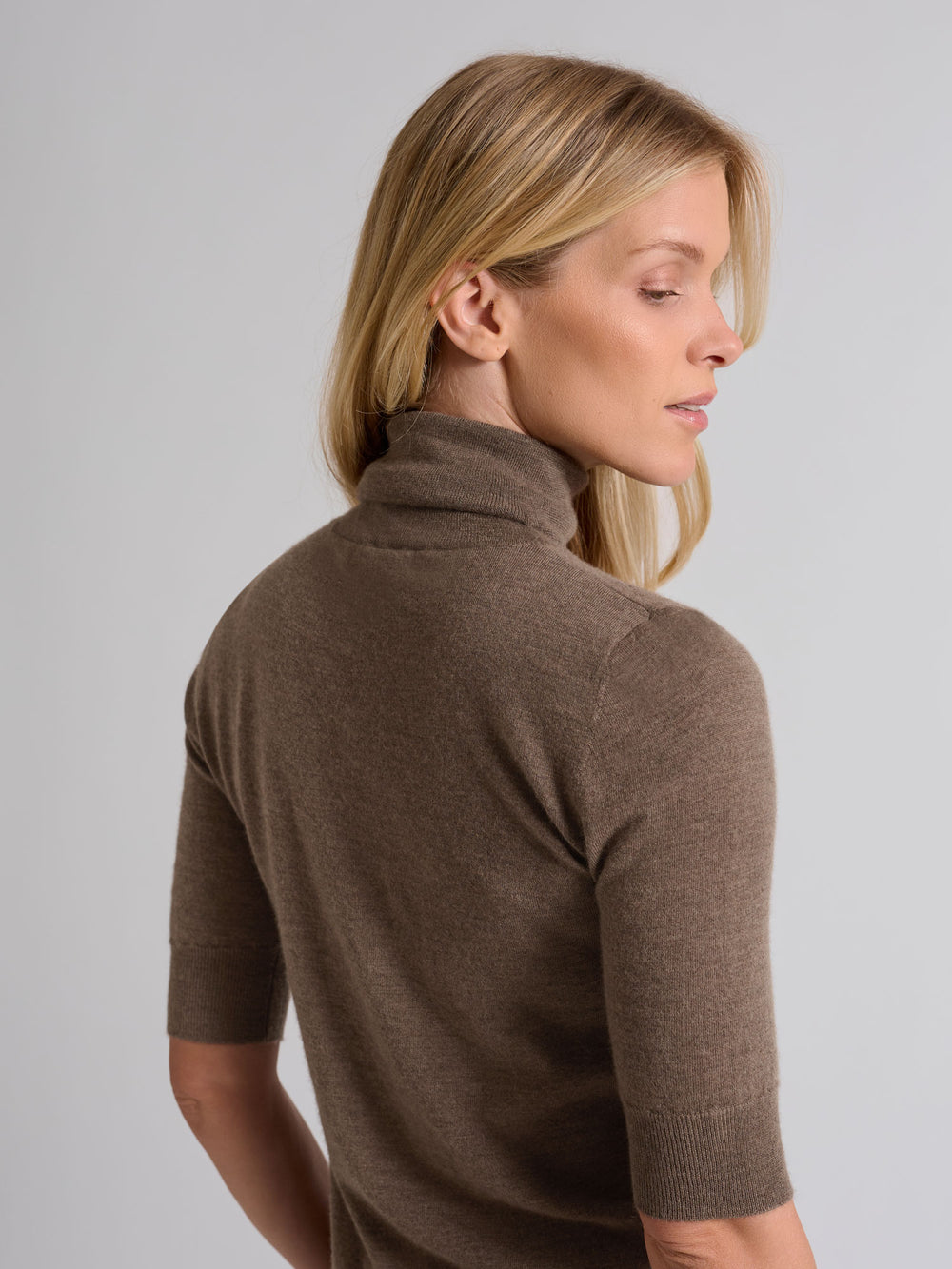 Woman wearing a brown turtleneck sweater against a gray background. Turtleneck cashmere t-shirt "Agnes" in 100% pure cashmere. Scandinavian design by Kashina. Color: Dark Toast.