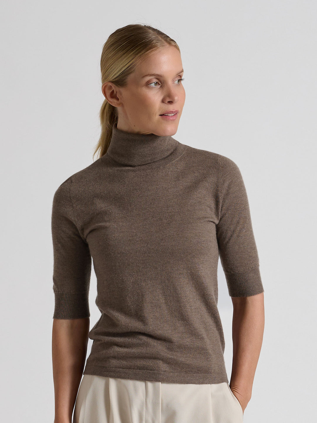 Woman wearing a brown turtleneck sweater against a gray background. Turtleneck cashmere t-shirt "Agnes" in 100% pure cashmere. Scandinavian design by Kashina. Color: Dark Toast.
