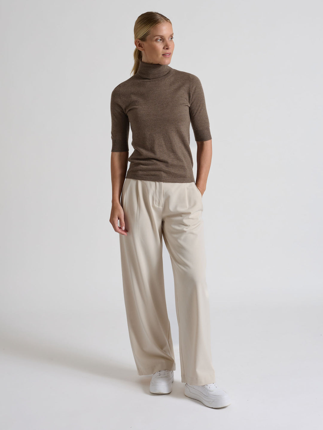 Woman wearing a brown turtleneck sweater against a gray background. Turtleneck cashmere t-shirt "Agnes" in 100% pure cashmere. Scandinavian design by Kashina. Color: Dark Toast.