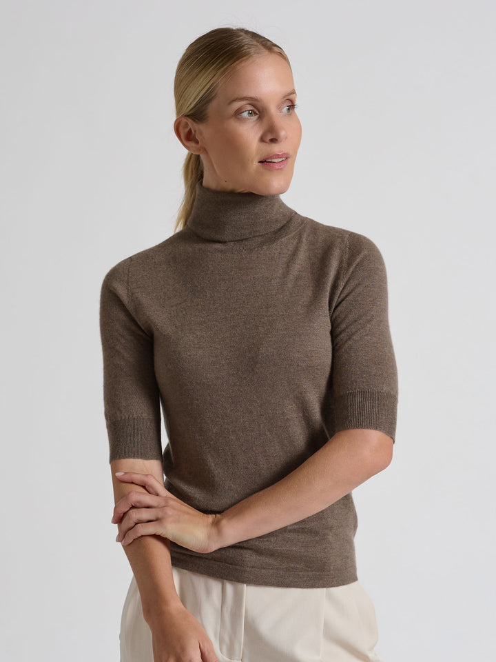 Woman wearing a brown turtleneck sweater against a gray background. Turtleneck cashmere t-shirt "Agnes" in 100% pure cashmere. Scandinavian design by Kashina. Color: Dark Toast.