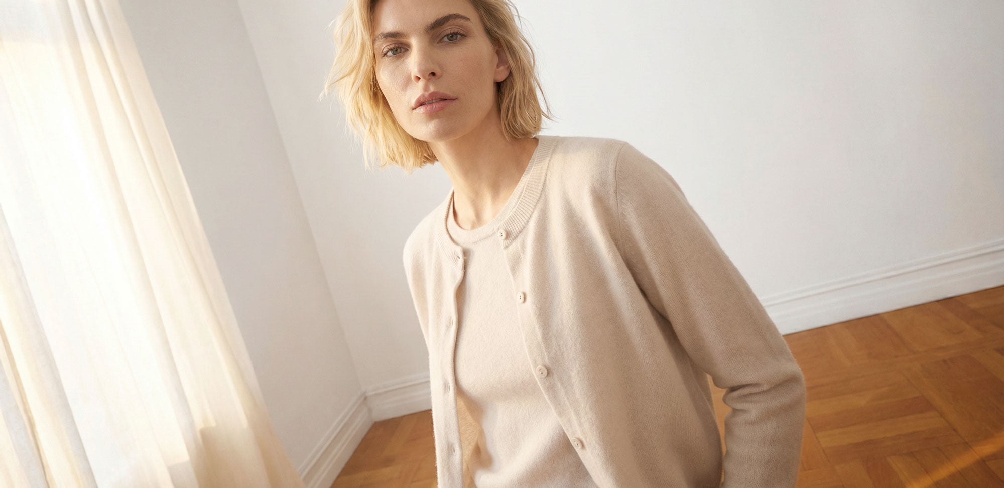Woman wearing a creme cashmere cardigan and creme cashmere shirt in a bright room with wooden floor and sheer curtains.